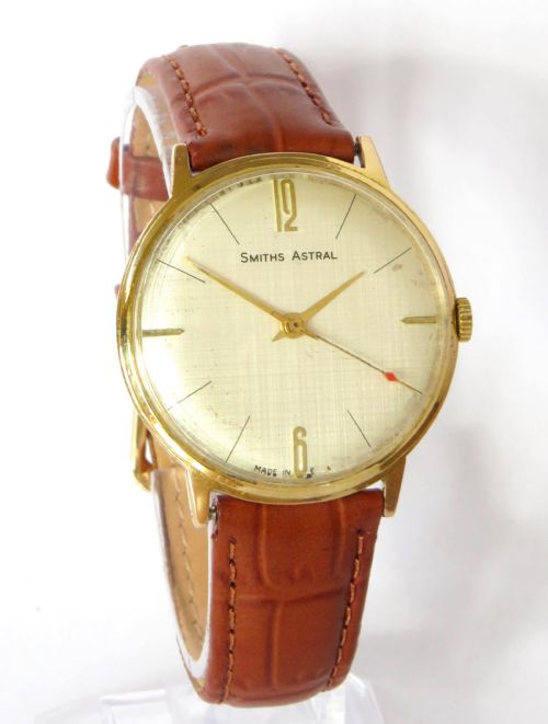 fig 1. A 1960s gents Smiths Astral wristwatch featuring a circular gold-plated case, textured cream dial, gold baton indices, and a brown leather strap.