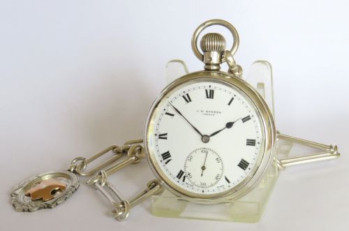 fig 1. A silver open-face pocket watch displaying black Roman numerals on a white dial, shown alongside a silver chain and decorative fob medallion.