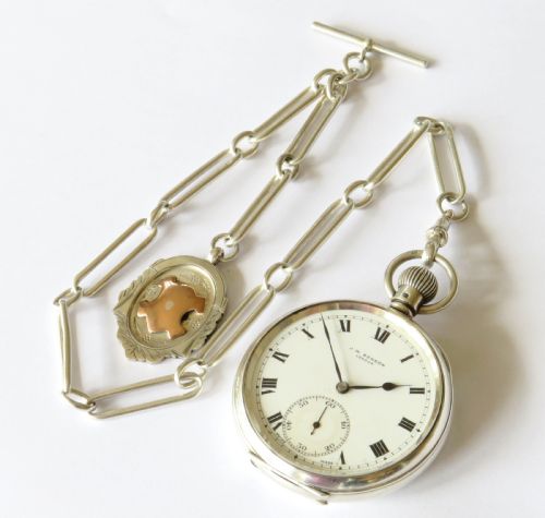 fig 2. A silver open-face pocket watch displaying black Roman numerals on a white dial, shown alongside a silver chain and decorative fob medallion.