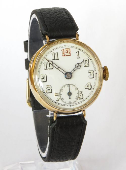 fig 1. An antique 9ct gold trench watch featuring a white enamel dial with black numerals, a red twelve, fixed wire lugs, and a black leather strap.