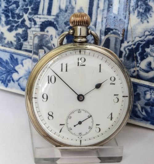 fig 1. An open-face antique silver pocket watch featuring a white enamel dial with black Arabic numerals, a sub-seconds dial, and a ribbed gold-toned crown.