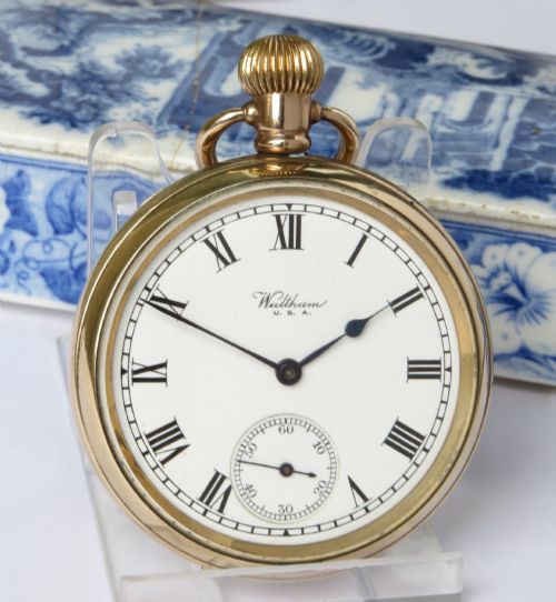 fig 1. A gold-toned open-face Waltham pocket watch featuring a white enamel dial with black Roman numerals and a sub-seconds dial.