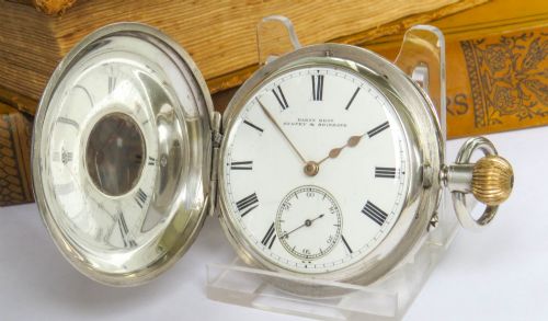 fig 1. An antique sterling silver half hunter pocket watch featuring an engine-turned case with blue enamel Roman numerals around a central aperture.