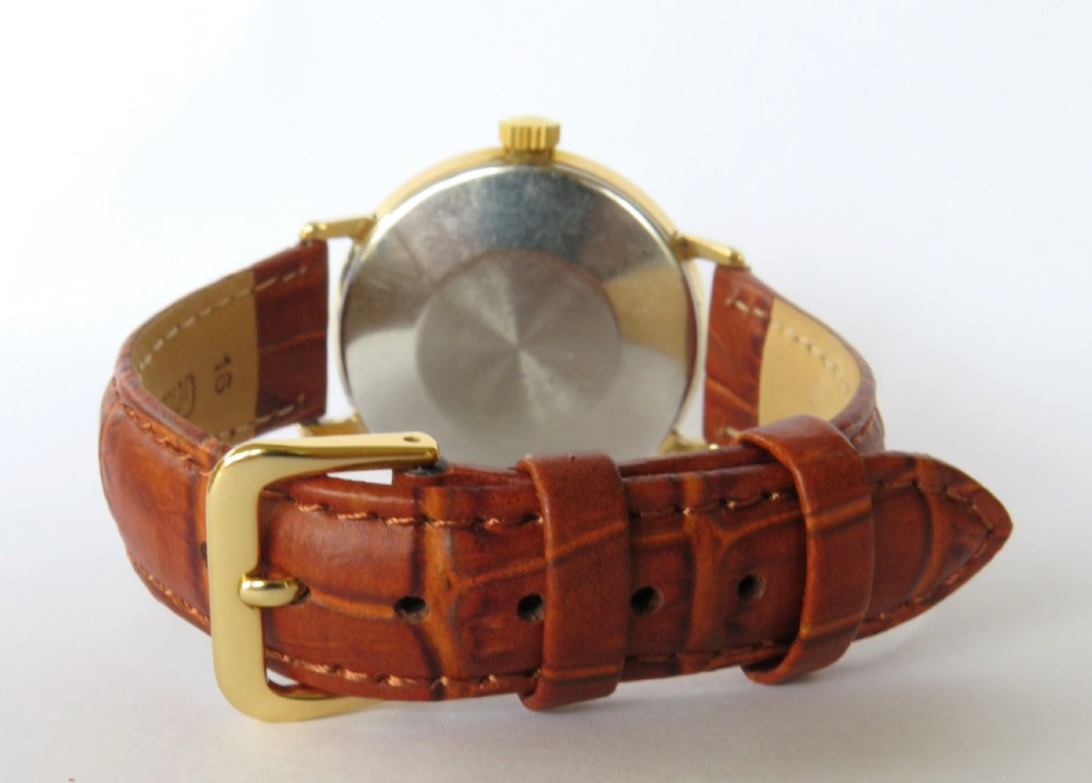 A 1960s Smiths Astral wristwatch featuring a gold-plated case, a cream dial with Arabic numerals at twelve, three, six, and nine, and a brown leather strap. View V