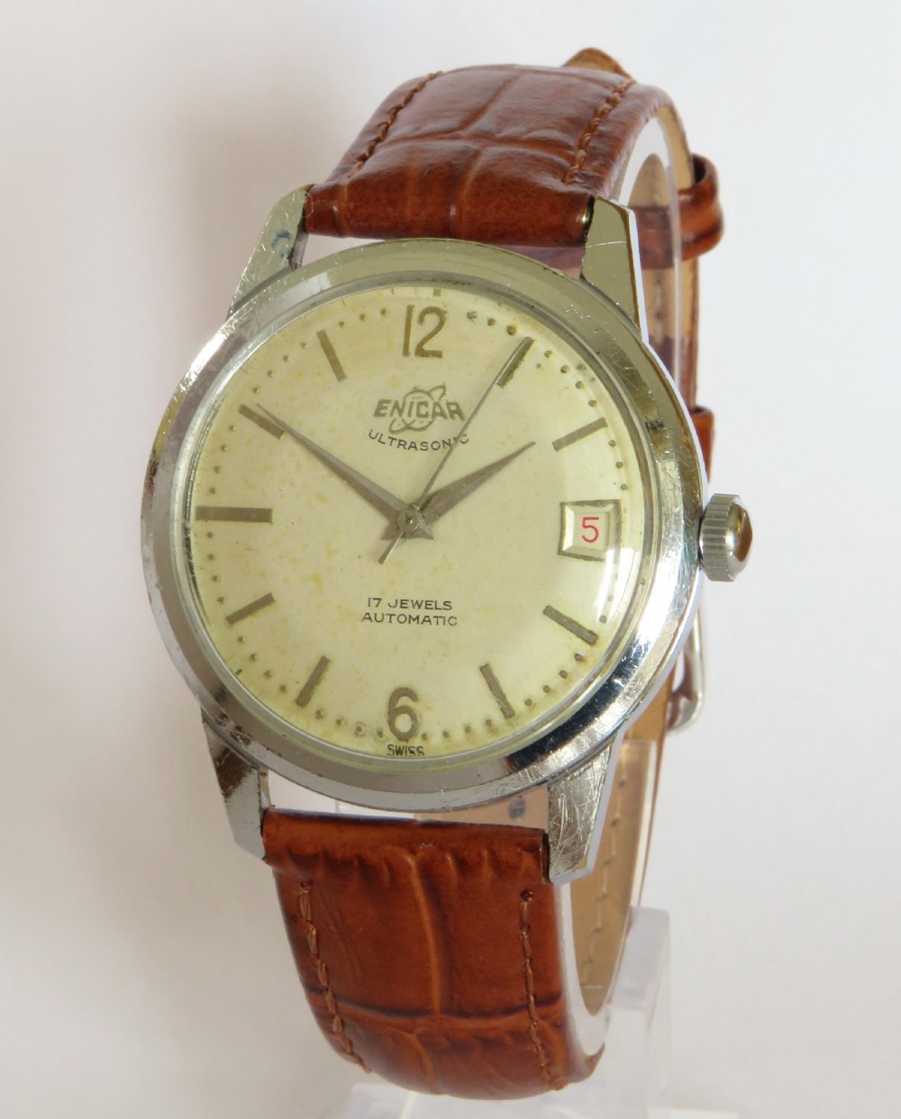 A 1950s mens Enicar Ultrasonic automatic wristwatch featuring a circular silver-tone case, cream dial with hour batons, date window, and a brown leather strap.