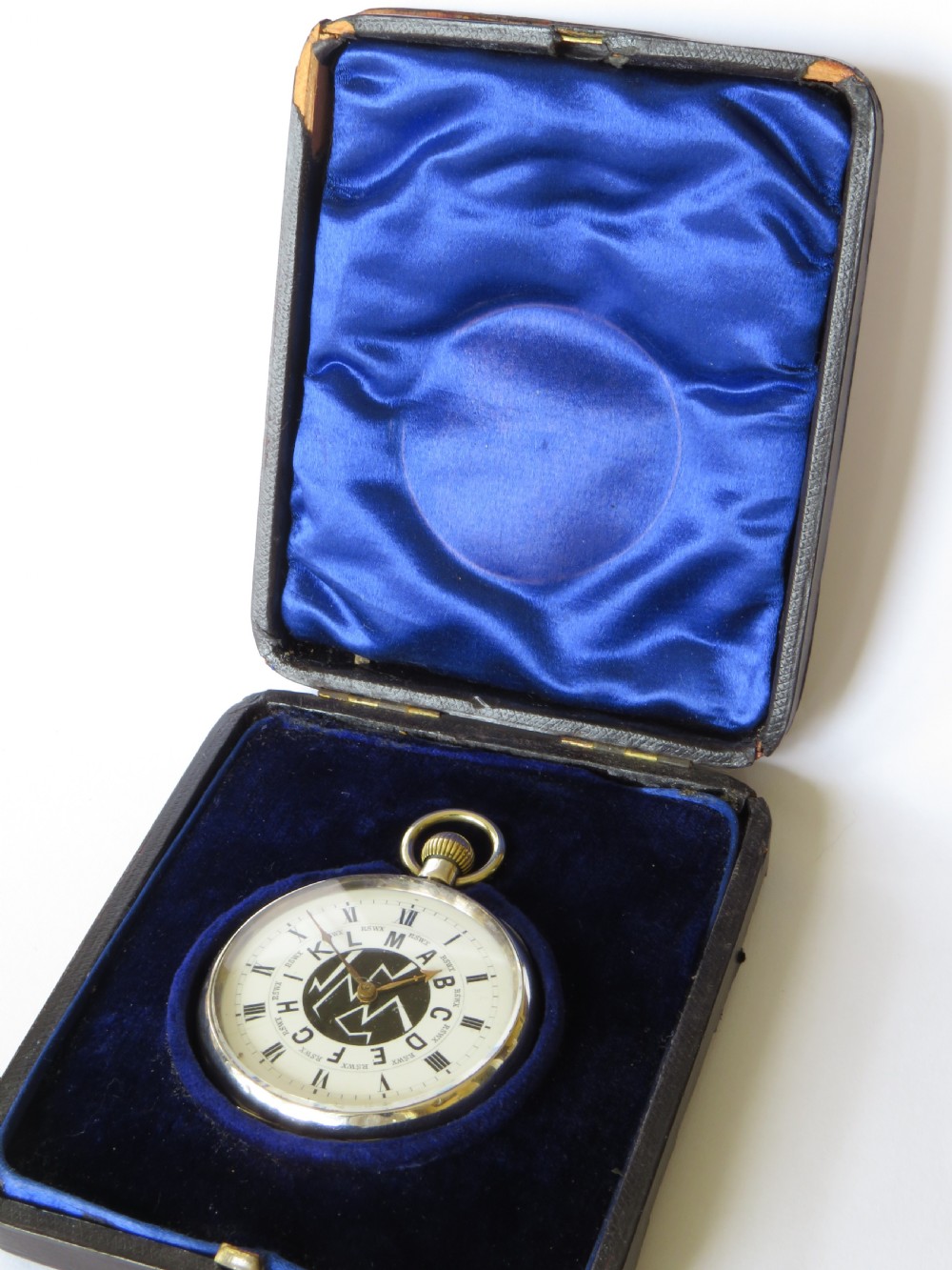 An open-face silver pocket watch featuring a white enamel dial with Roman numerals, central telegraphic letter codes, and gold-tone hands. View III
