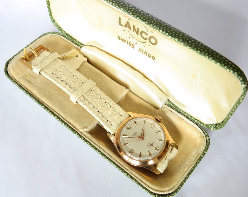 A 1950s gold-plated Lanco mechanical wrist watch with a cream dial and light leather strap resting inside its original green patterned presentation case.