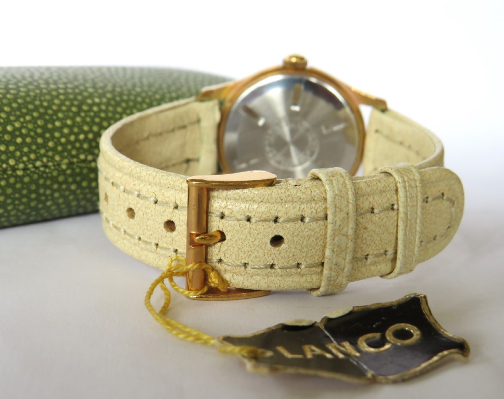 A 1950s gold-plated Lanco mechanical wrist watch with a cream dial and light leather strap resting inside its original green patterned presentation case. View V