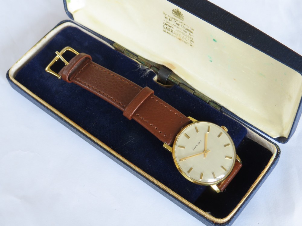 A 1960s gents Garrard wristwatch with a gold-toned case, off-white dial, baton markers, and brown leather strap resting inside a navy blue velvet presentation box.