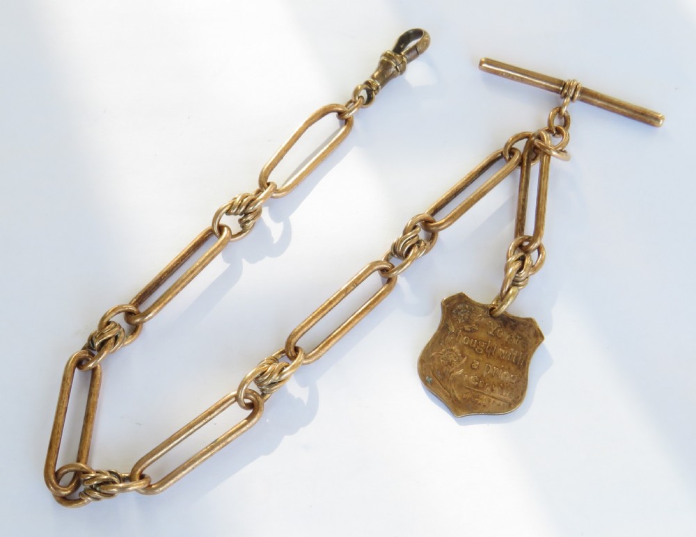 An antique rolled rose gold pocket watch chain featuring elongated open links, a T-bar, swivel clasp, and an engraved shield-shaped fob.