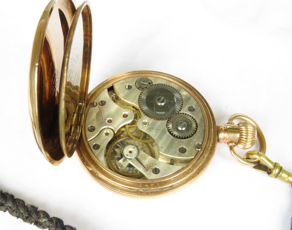 An open-face gold pocket watch featuring a white dial with Roman numerals and an accompanying black braided strap with a brass T-bar. View III