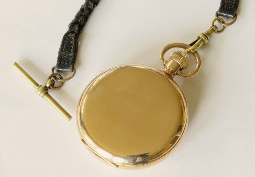 An open-face gold pocket watch featuring a white dial with Roman numerals and an accompanying black braided strap with a brass T-bar. View IV