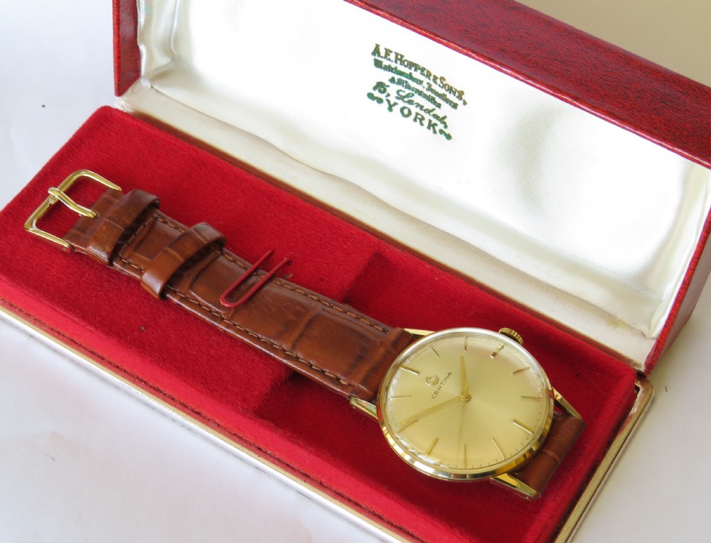 A 1960s Certina gents wristwatch featuring a gold dial, baton hour markers, and a brown leather strap alongside its red presentation box. View II
