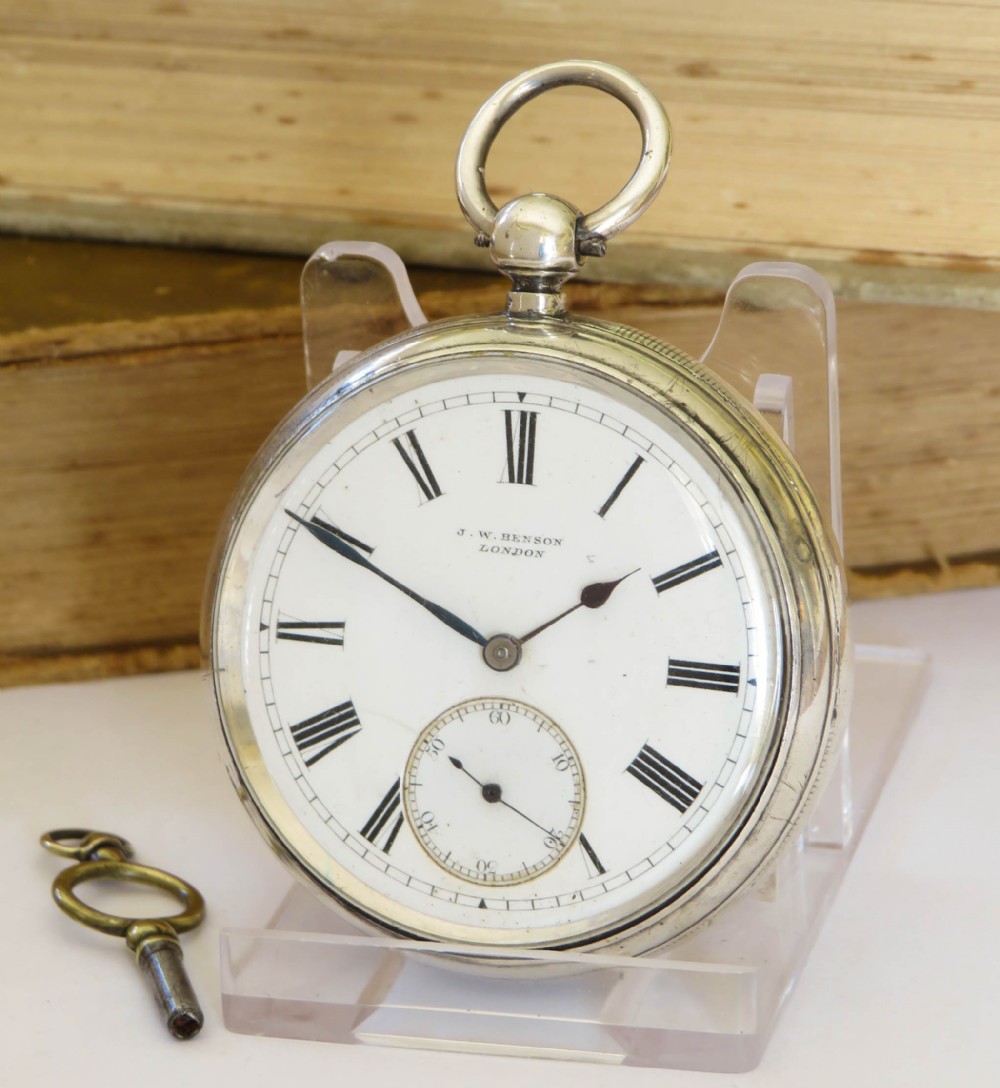 An open-face silver pocket watch with a white enamel dial, black Roman numerals, blued steel hands, and a separate winding key.