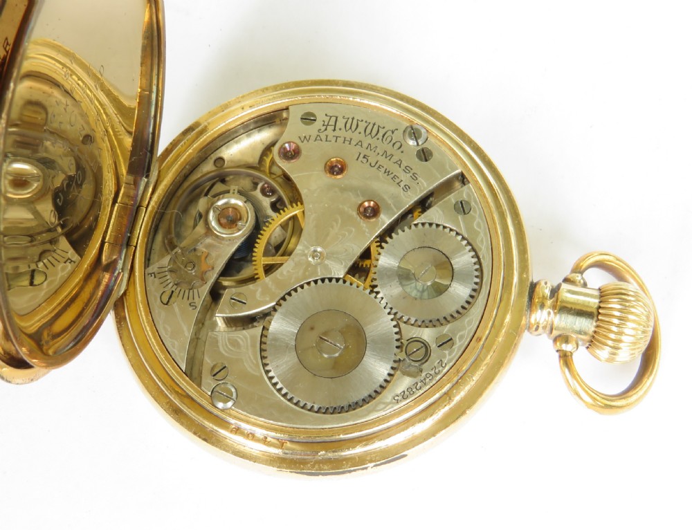 A gold-toned antique Waltham half hunter pocket watch featuring a central glass window surrounded by black Roman numerals on the outer casing. View VI