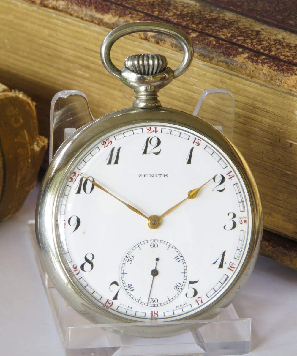 A 1939 Zenith open-face pocket watch featuring a white enamel dial with black Arabic numerals, secondary red 24-hour markers, a subsidiary seconds dial, and a metallic silver case.