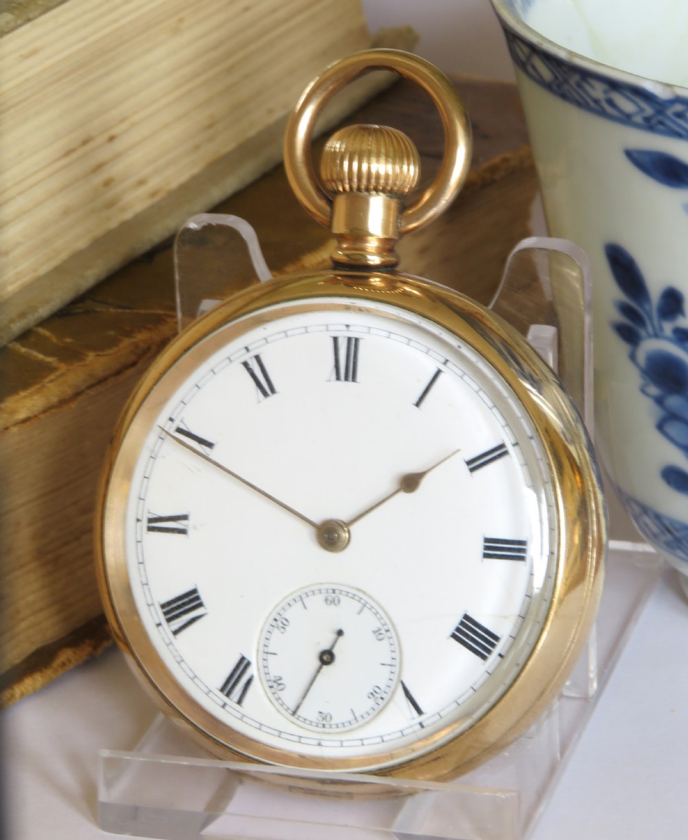An open-face gold pocket watch featuring a white enamel dial, black Roman numerals, and a subsidiary seconds dial at six oclock.