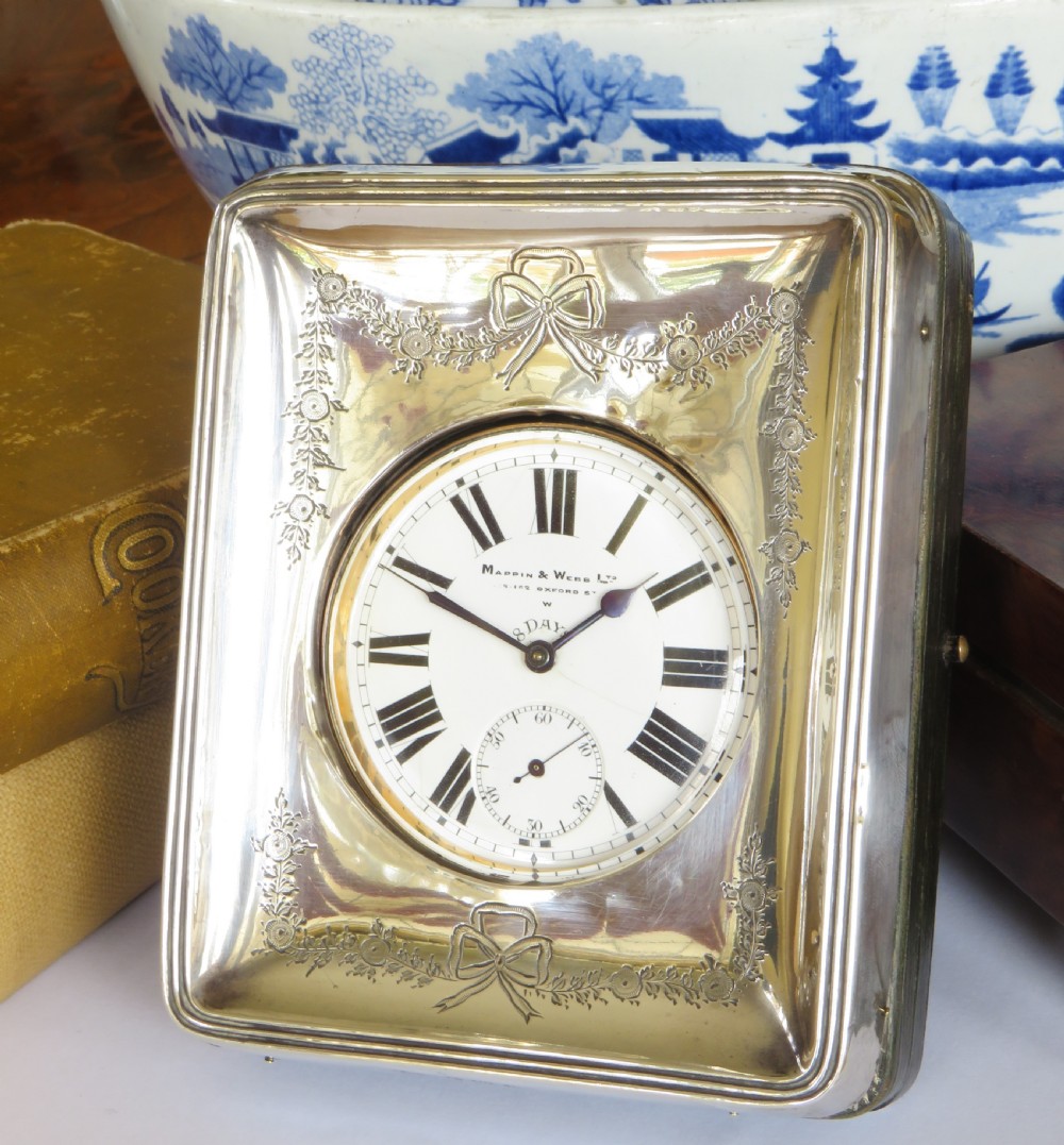 A rectangular silver travel desk clock featuring bow and garland engravings surrounding a white enamel dial with Roman numerals.
