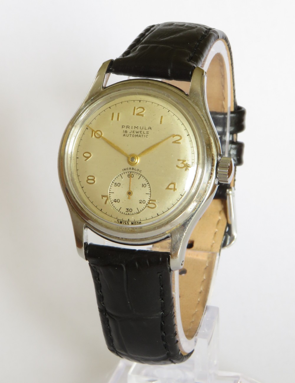 A 1950s gentlemens Primula Swiss automatic wristwatch featuring a silver-tone case, cream dial with gold-tone Arabic numerals, sub-seconds dial, and a black leather strap.
