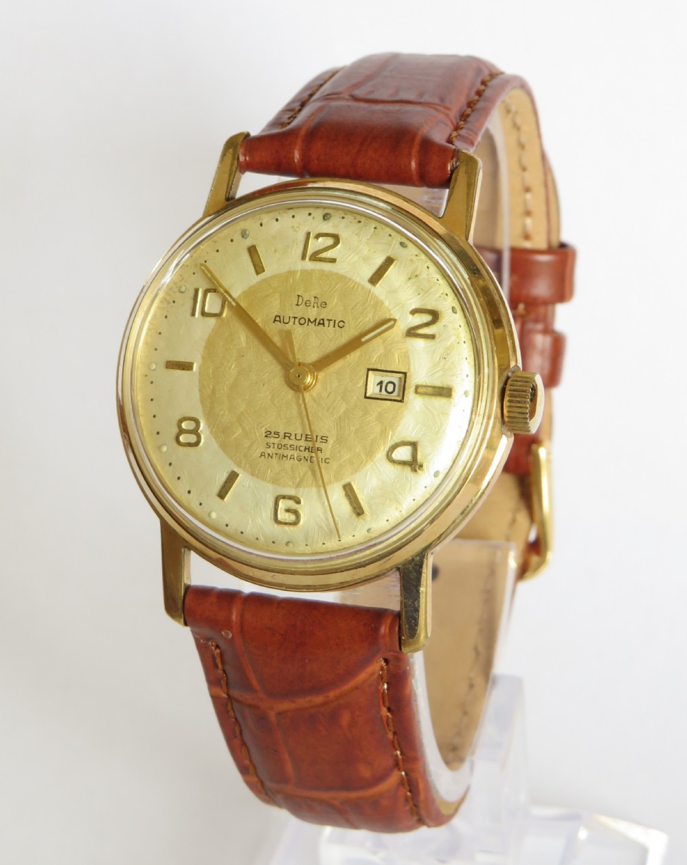 A 1950s gents gold-toned automatic wristwatch featuring a textured dial, Arabic numerals, a date window at 3 oclock, and a brown leather strap.