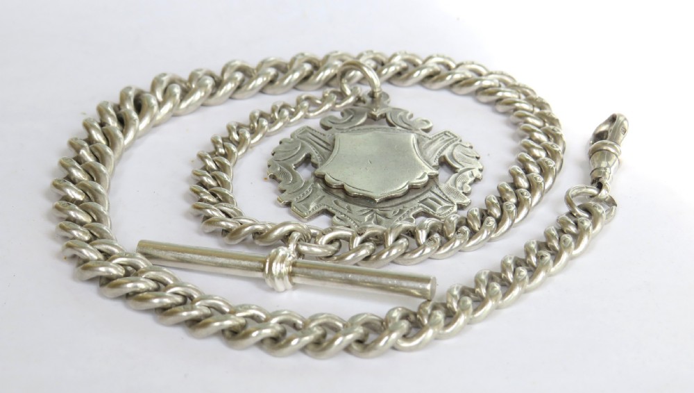 A heavy silver Albert pocket watch chain laid in a circular pattern, featuring curb links, a T-bar attachment, swivel clasp, and a central shield-shaped fob medallion.