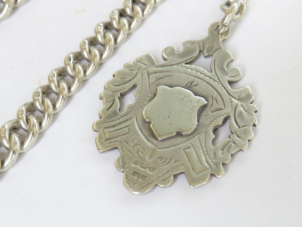 A heavy silver Albert pocket watch chain laid in a circular pattern, featuring curb links, a T-bar attachment, swivel clasp, and a central shield-shaped fob medallion. View IV