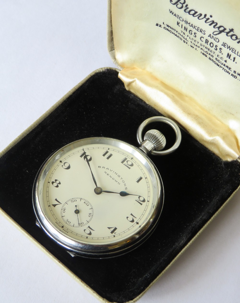 A 1950s Bravingtons Renown open-face pocket watch with a cream dial, black Arabic numerals, and blued steel hands, set inside its cream presentation case. View II