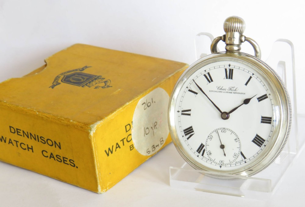 An antique 1922 open-face silver pocket watch featuring a white enamel dial with Roman numerals alongside its original yellow Dennison box.
