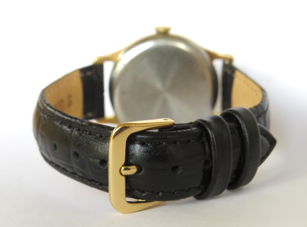 A 1950s circular gold-plated wristwatch featuring a cream dial, black Roman numerals, subsidiary seconds dial, and a black leather strap. View V