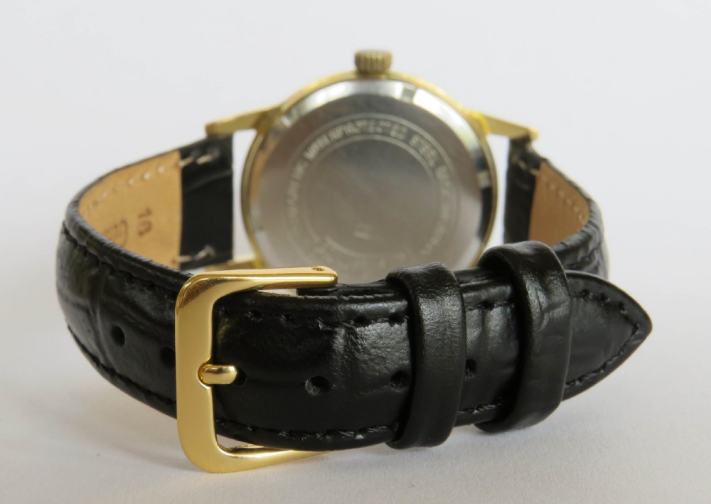 A 1950s gents Heloisa wrist watch with a circular gold-tone case, cream dial with gold baton markers, date aperture at three oclock, and black leather strap. View V