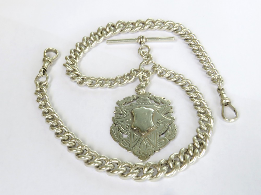 An antique silver double Albert pocket watch chain featuring curb links, two swivel dog clips, a central T-bar, and a decorative shield-shaped pendant fob.