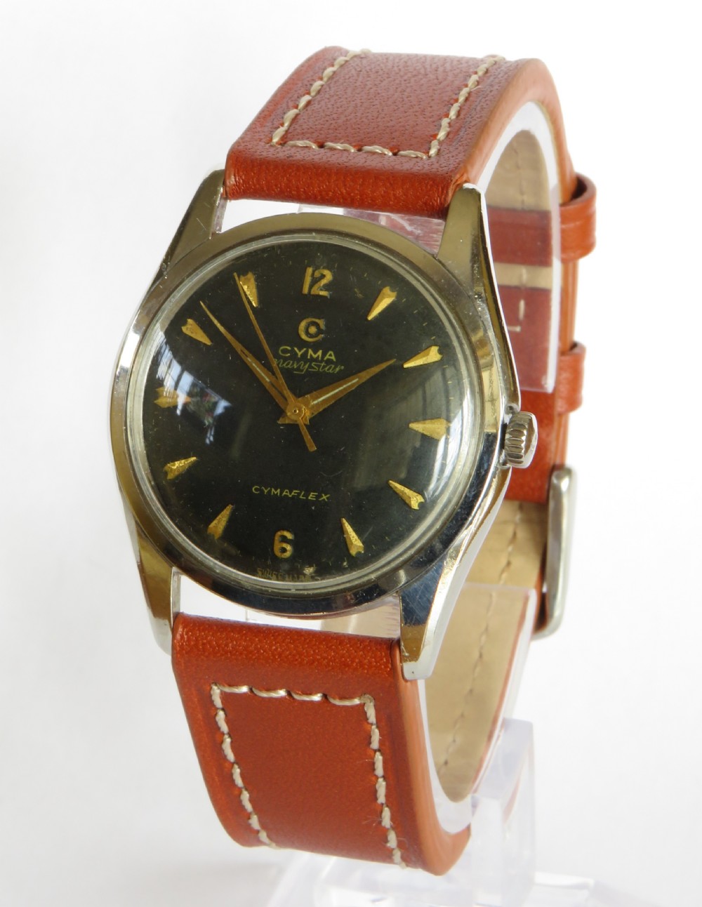 A 1960s Cyma Navystar wristwatch featuring a black dial with gold-toned markers and hands, housed in a polished metal case on a tan leather strap.