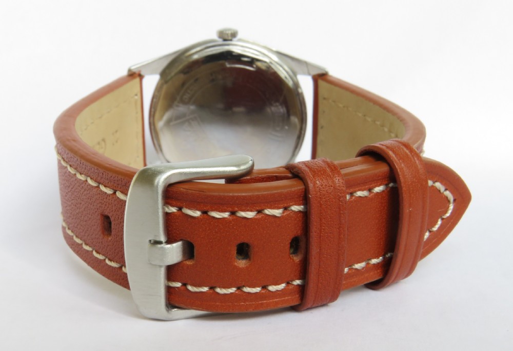 A 1960s Cyma Navystar wristwatch featuring a black dial with gold-toned markers and hands, housed in a polished metal case on a tan leather strap. View IV