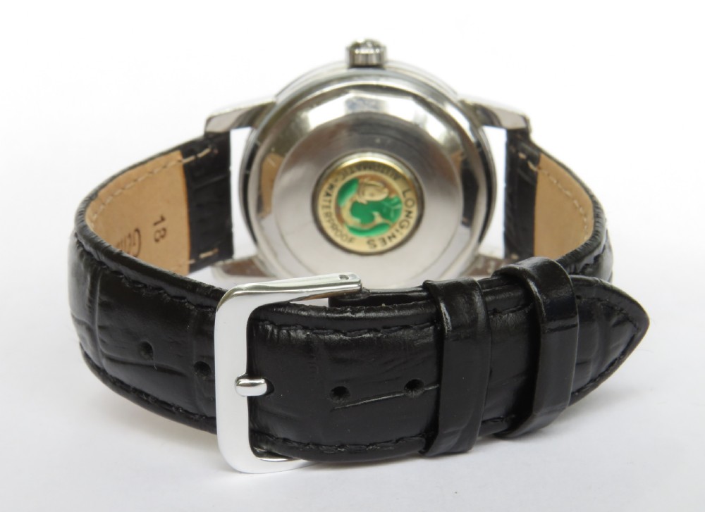 A 1957 Longines Conquest automatic wristwatch featuring a stainless steel case, patinated cream dial, gold-tone hour markers, and a black leather strap. View V