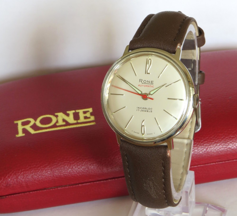 A 1960s mens Rone automatic wristwatch featuring a champagne dial, gold-tone casing, and a brown leather strap.