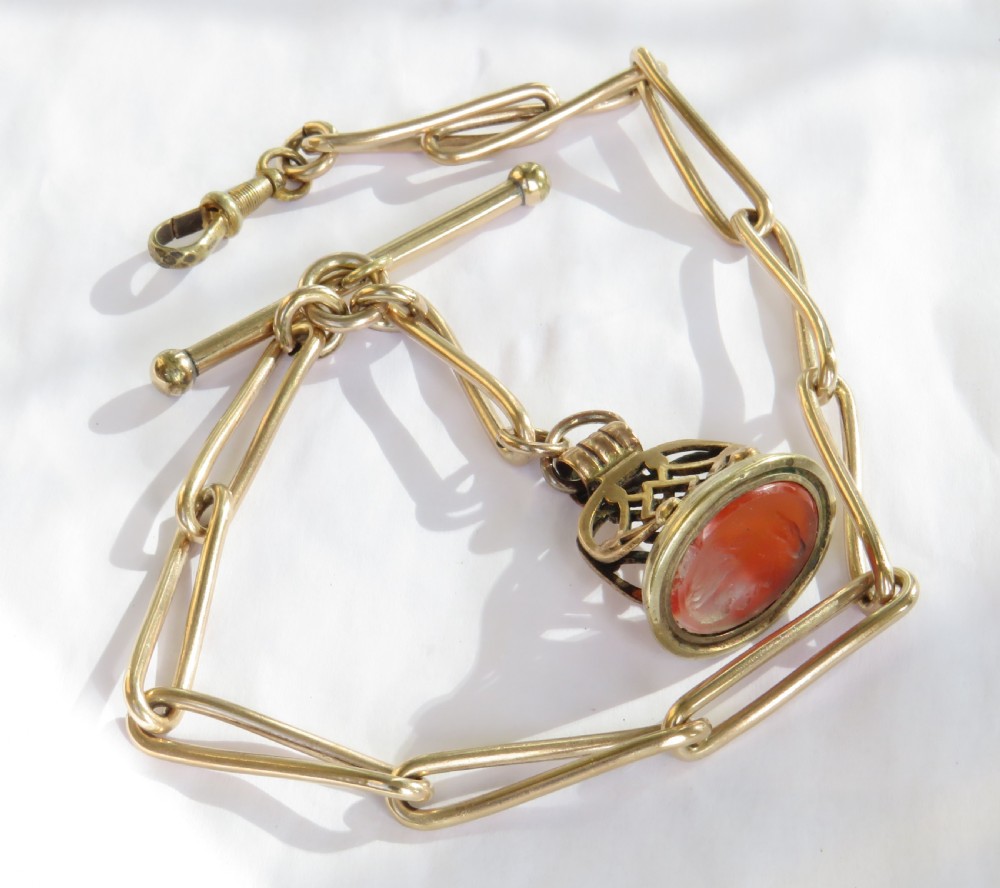 An antique 1920s rolled rose gold watch Albert chain featuring elongated trombone links, a T-bar, swivel clasp, and a carved carnelian fob pendant.