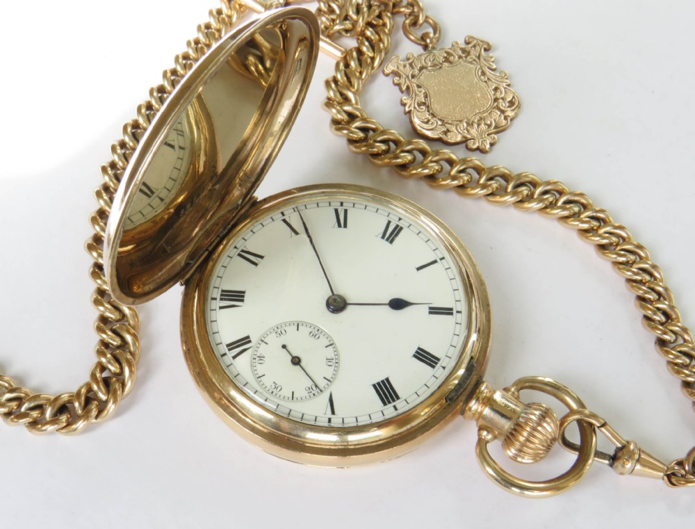 An antique gold-coloured full hunter pocket watch open to reveal a white dial with Roman numerals, alongside a curb link chain and decorative shield fob.