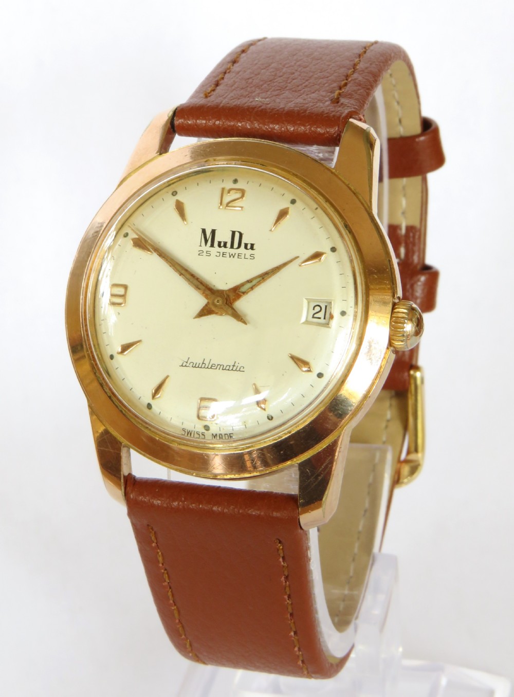 A 1950s gents MuDu Doublematic wristwatch featuring a gold-toned case, cream dial with gold hour markers, date aperture at three oclock, and a brown leather strap.