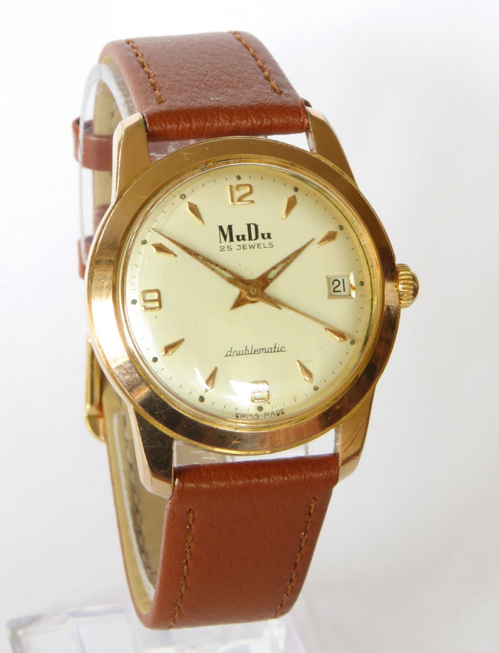 A 1950s gents MuDu Doublematic wristwatch featuring a gold-toned case, cream dial with gold hour markers, date aperture at three oclock, and a brown leather strap. View II