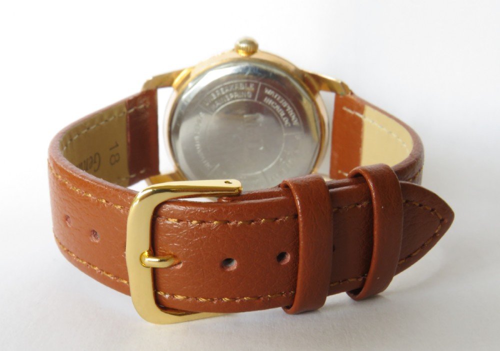 A 1950s gents MuDu Doublematic wristwatch featuring a gold-toned case, cream dial with gold hour markers, date aperture at three oclock, and a brown leather strap. View V