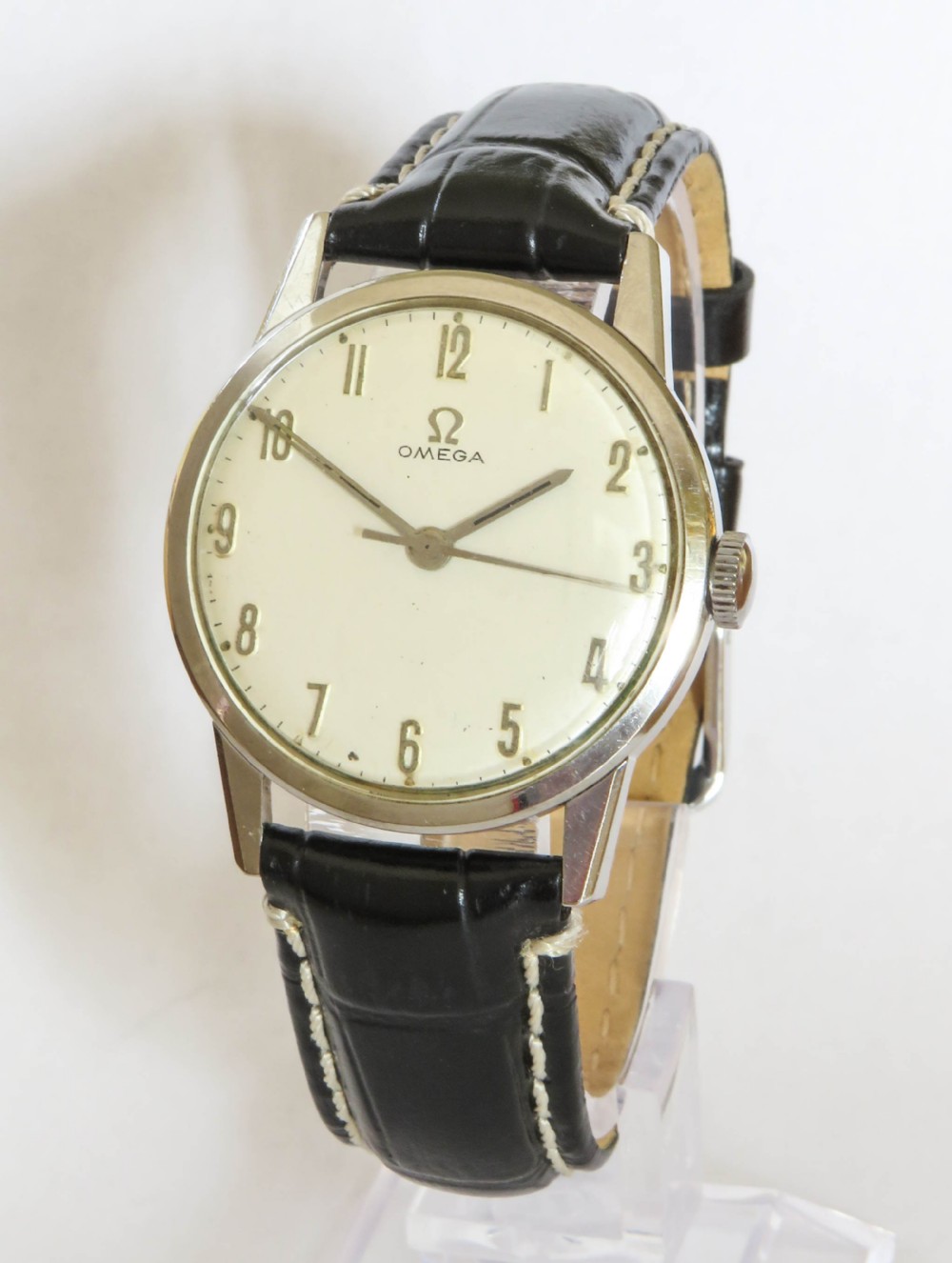 A 1963 mens Omega wrist watch featuring a round stainless steel case, a cream dial with Arabic numerals, and a black leather strap with white stitching.