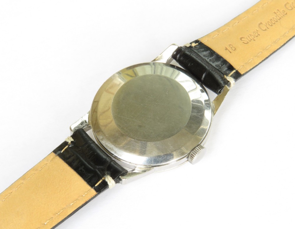 A 1963 mens Omega wrist watch featuring a round stainless steel case, a cream dial with Arabic numerals, and a black leather strap with white stitching. View V