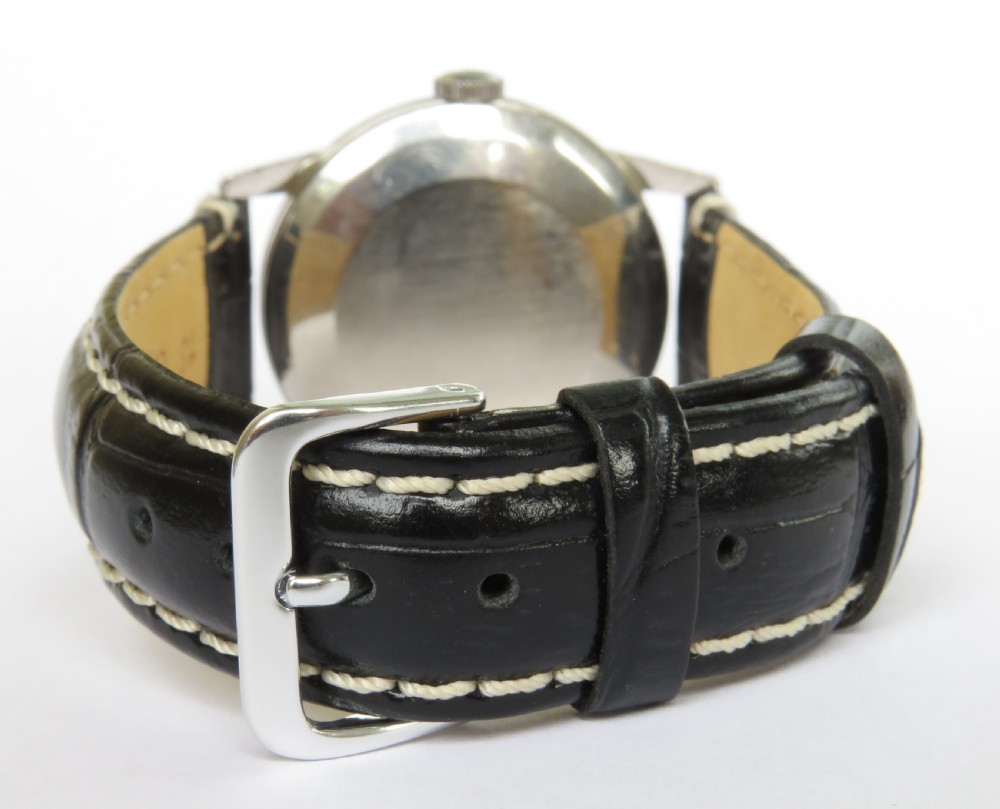 A 1963 mens Omega wrist watch featuring a round stainless steel case, a cream dial with Arabic numerals, and a black leather strap with white stitching. View VI