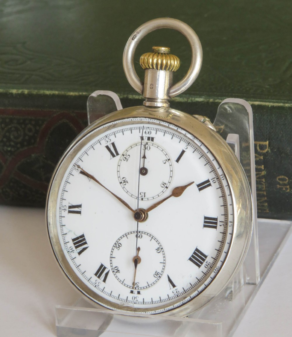 An open-face antique silver chronograph pocket watch featuring a white enamel dial with black Roman numerals, two subsidiary dials, and a gold-toned winding crown.