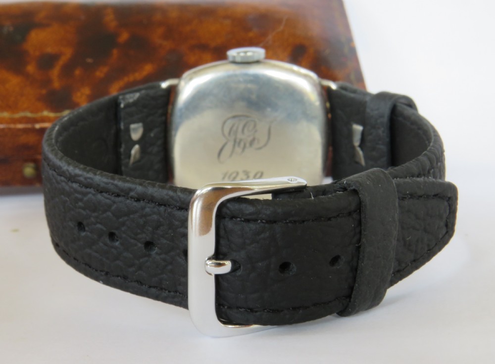 An antique 1920s cushion-shaped sterling silver wristwatch with a white enamel dial, black Arabic numerals, a red twelve, and a black leather strap, presented in an open fitted velvet box. View VI