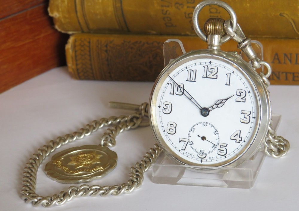 An open-face silver pocket watch with a white enamel dial, black numerals, and sub-seconds dial, attached to a silver curb-link chain with a military badge fob.