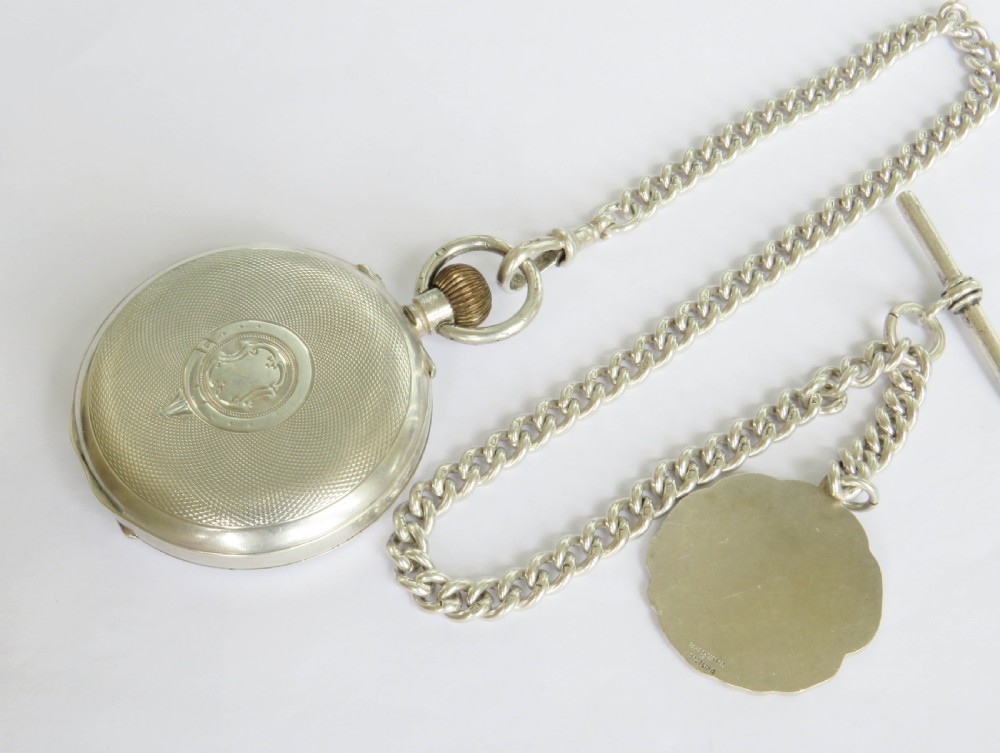 An open-face silver pocket watch with a white enamel dial, black numerals, and sub-seconds dial, attached to a silver curb-link chain with a military badge fob. View VI