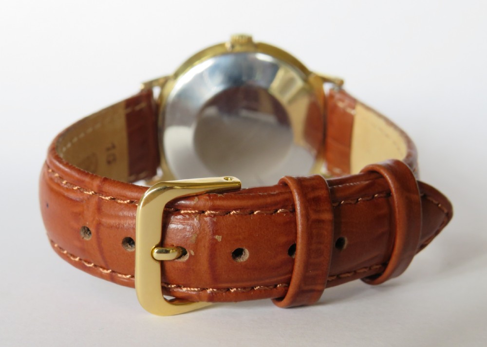 A 1960s gents Smiths Astral wristwatch featuring a circular gold-plated case, textured cream dial, gold baton indices, and a brown leather strap. View V