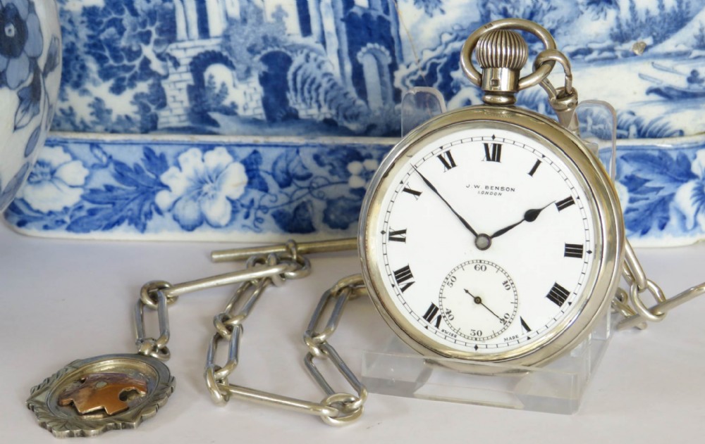 A silver open-face pocket watch displaying black Roman numerals on a white dial, shown alongside a silver chain and decorative fob medallion.