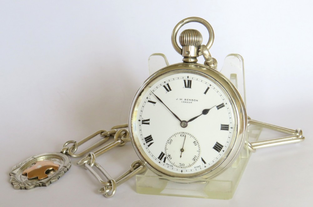 A silver open-face pocket watch displaying black Roman numerals on a white dial, shown alongside a silver chain and decorative fob medallion. View II